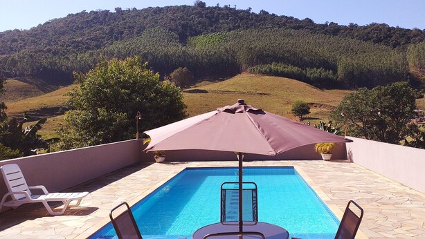 A rectangular pool with a clear blue water surface, surrounded by a tiled deck. There are two white lounge chairs and a large umbrella providing shade. The deck is bordered by a low wall, and the background features a hilly landscape with dense forest. - Chacara Recanto Paraíso is in a privileged location. (Serra Negra)