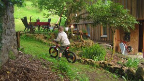 Un enfant fait du vélo sur un chemin de terre dans un jardin, avec une clôture en bois, un mur de pierre et un bâtiment en bois en arrière-plan.