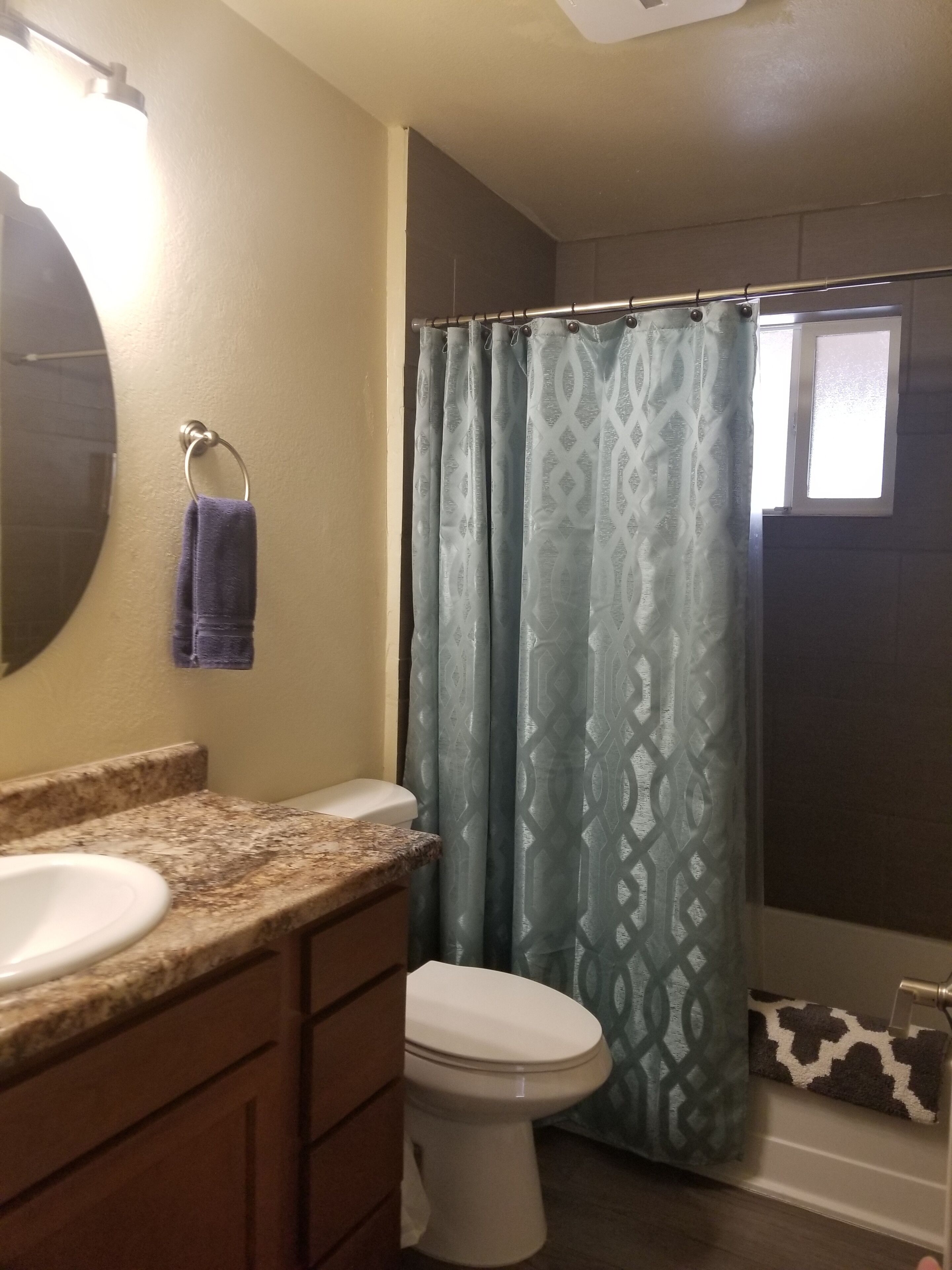 Une salle de bain avec un comptoir en granit, des toilettes blanches, une serviette suspendue à un anneau métallique et un rideau de douche à motifs.