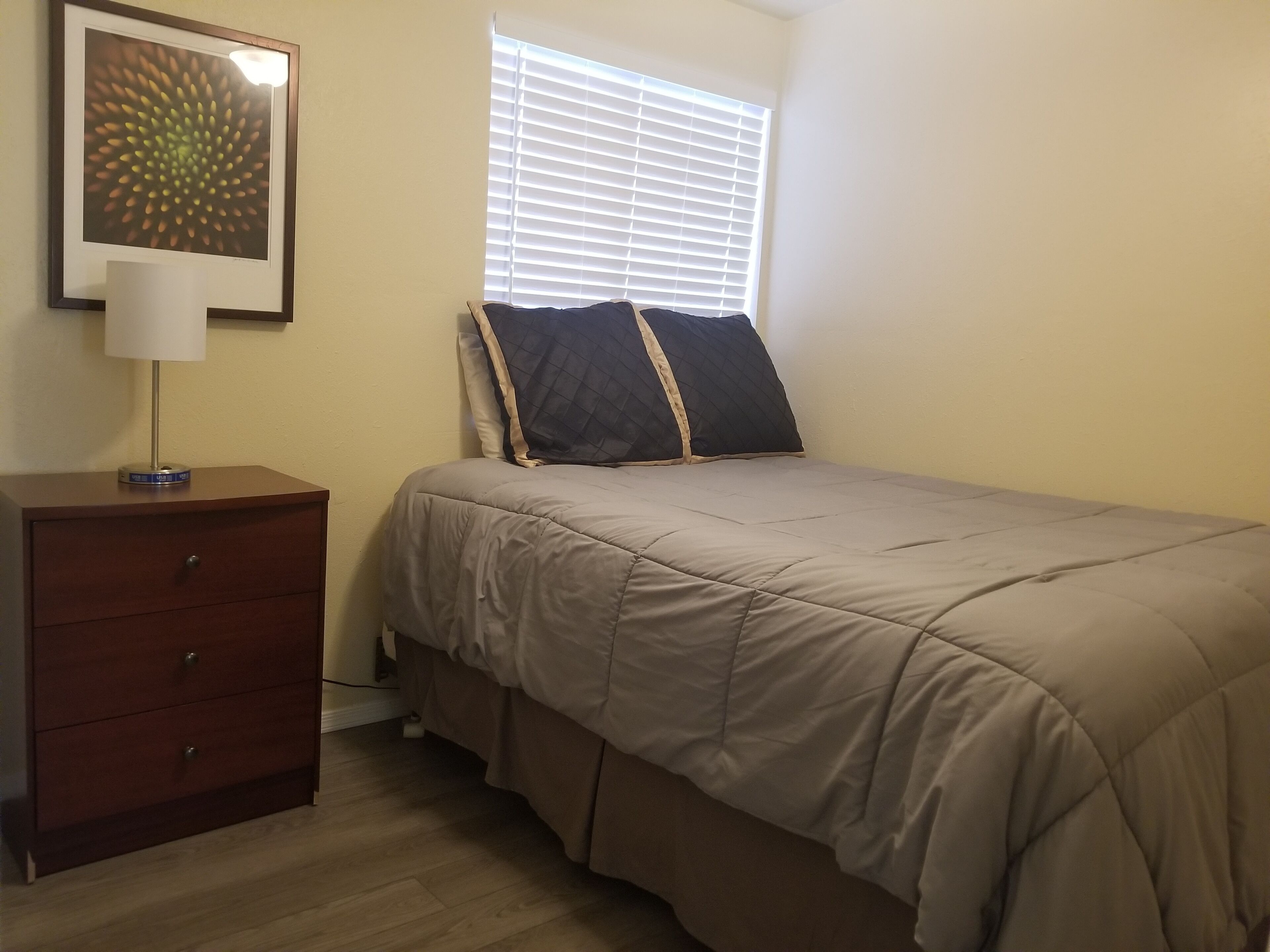 A bedroom with a bed, a nightstand, a lamp, a window with blinds, and a wall-mounted picture.
