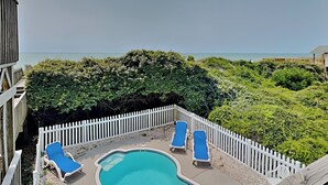 A pool area with two blue lounge chairs and a small pool, surrounded by a white picket fence and lush greenery. - Tipsy Turtle | Ocean Views + Pool! (North Topsail Beach)