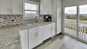 A modern kitchen with white cabinets, a granite countertop, a window with blinds, a coffee maker, and a view of the outdoors through a sliding glass door. - Tipsy Turtle | Ocean Views + Pool! (North Topsail Beach)