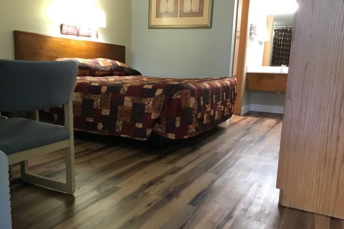 Una habitación con cama, una silla, suelo de madera y una ventana con cortinas.