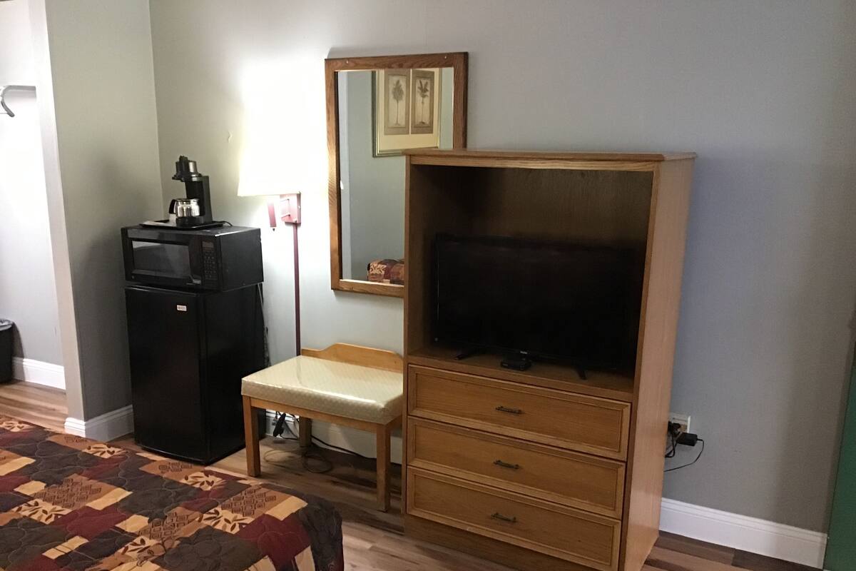 Un dormitorio con una cama, un aparador de madera con televisor, un escritorio pequeño y un refrigerador.