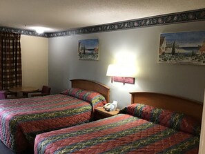 A hotel room with two beds, a small table, a lamp, and two paintings on the wall.