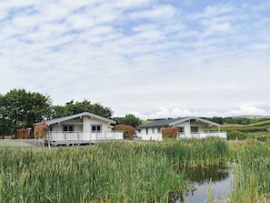 Two single-story buildings with white exteriors and balconies, situated by a calm body of water with tall green reeds.