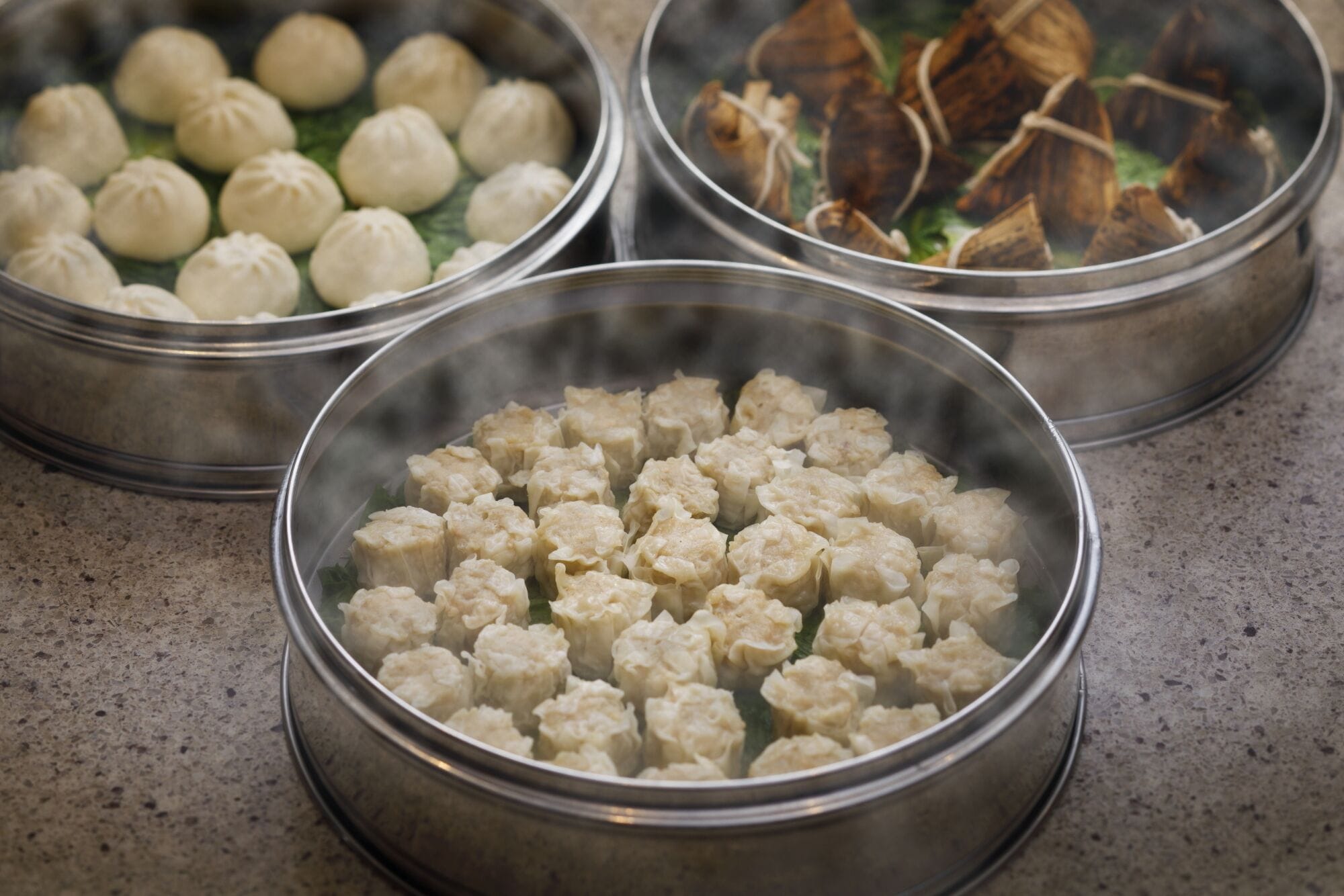 Three metal steamer baskets with food, one containing dumplings, another with a different type of dumpling, and the third with what appears to be meat and vegetables.