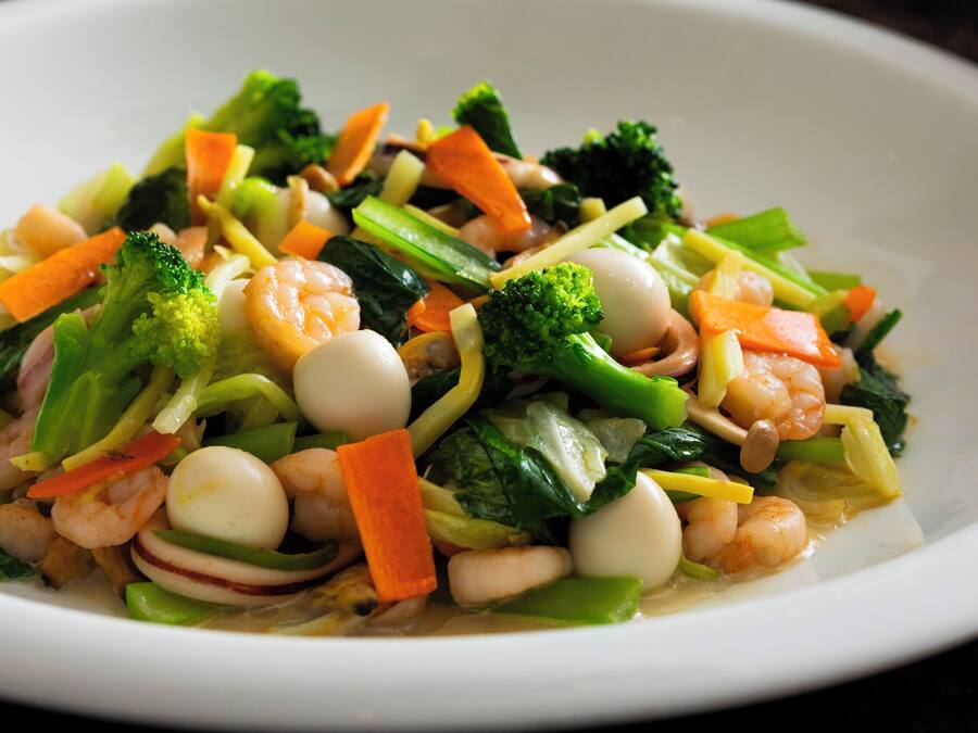 Une assiette blanche contenant un plat de légumes sautés, avec des crevettes, des brocolis, des carottes et d’autres légumes.