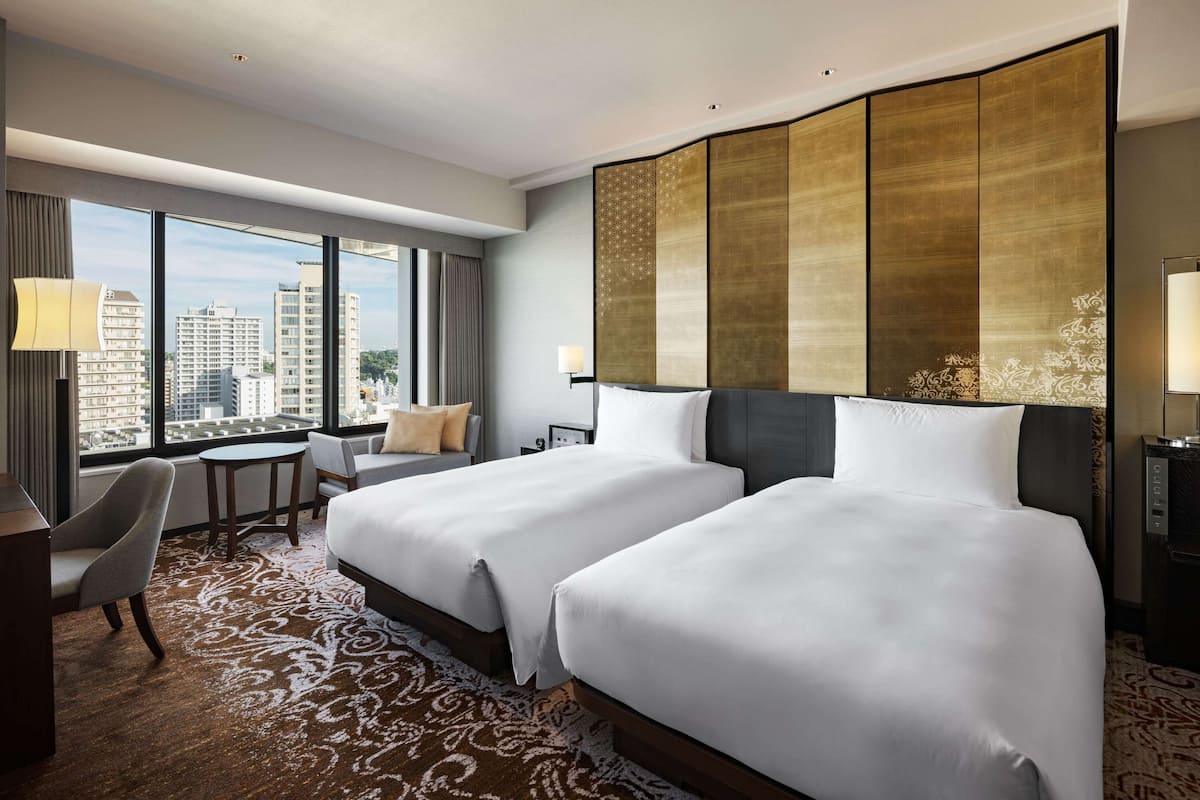 A hotel room with two beds, a desk, a chair, and a large window offering a city view.