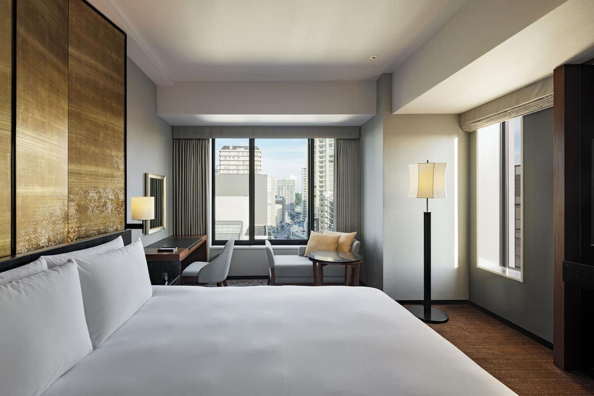 A hotel room with a large bed, a desk, a chair, a standing lamp, and a view of the cityscape.