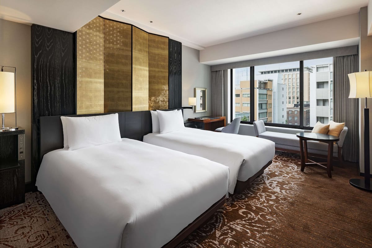 A hotel room with two large beds, a small table, a sofa, and a large window offering a city view.
