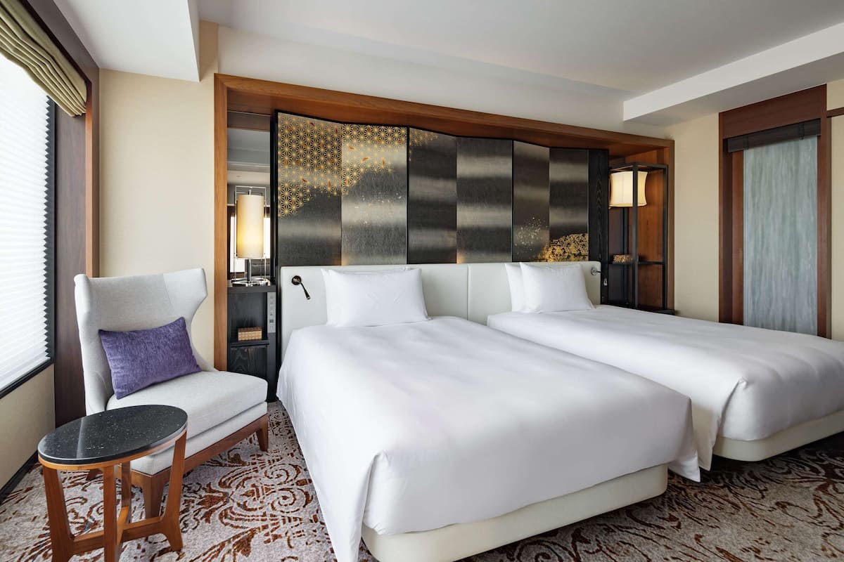 A hotel room with two beds, a chair, a small table, and a decorative headboard.