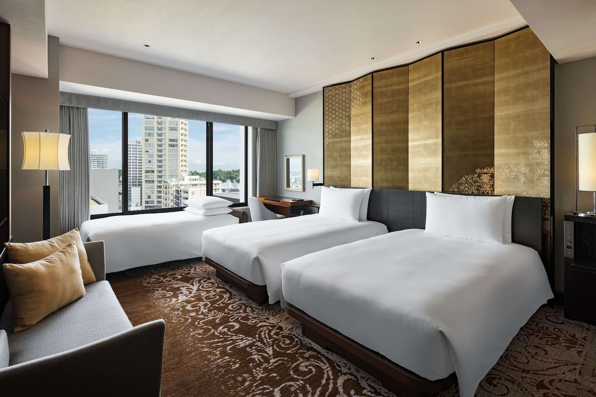 A hotel room with two beds, a sofa, a desk, and a large window offering a city view.