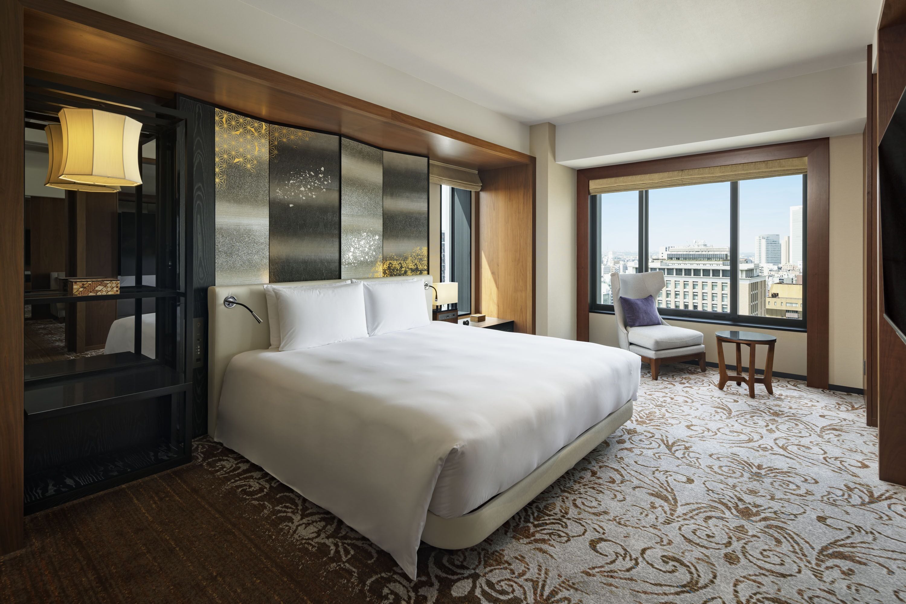 A hotel room with a large bed, a desk, a chair, a TV, and a view of the city.