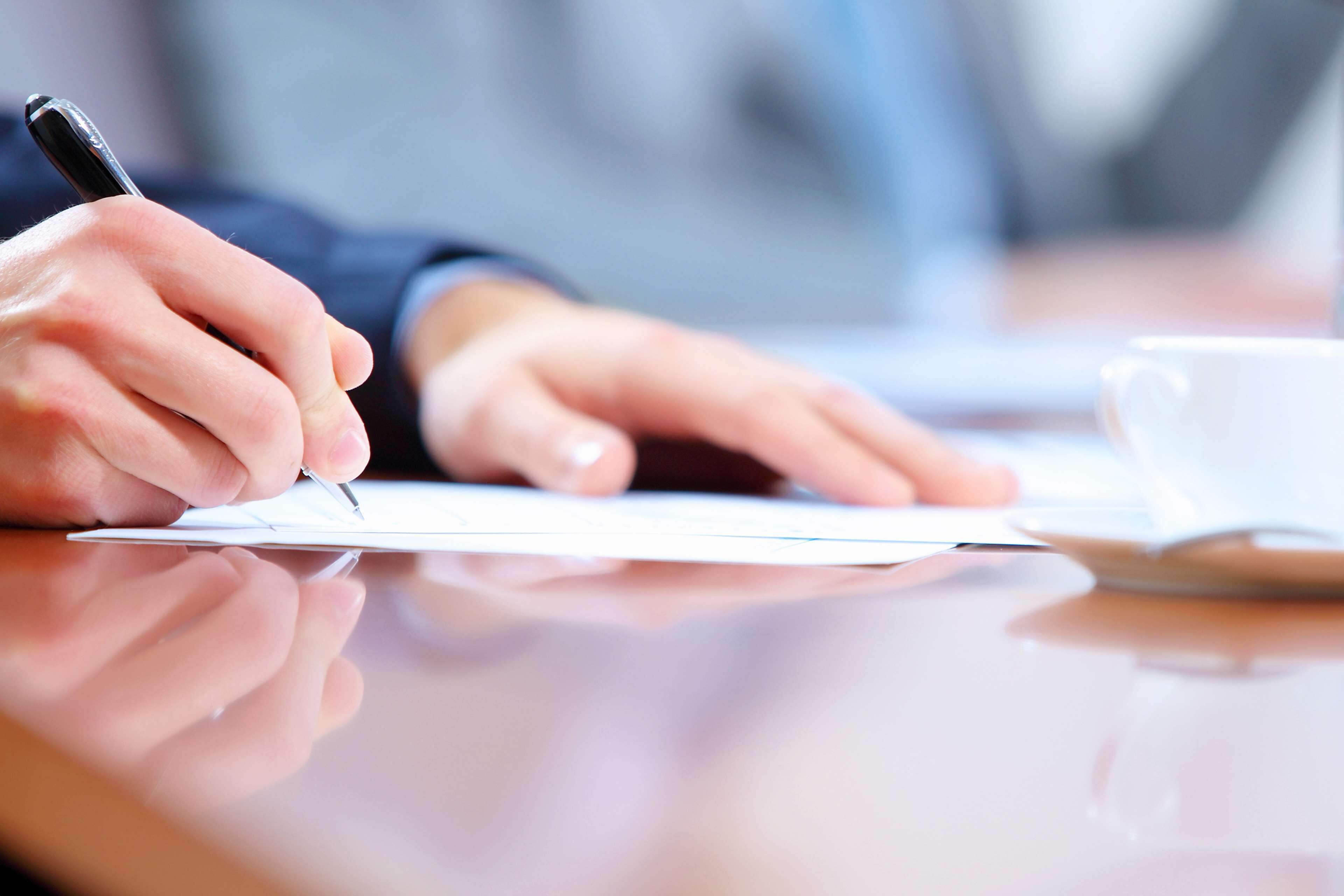 A person writing on a document with a pen, a cup, and a laptop on a table.