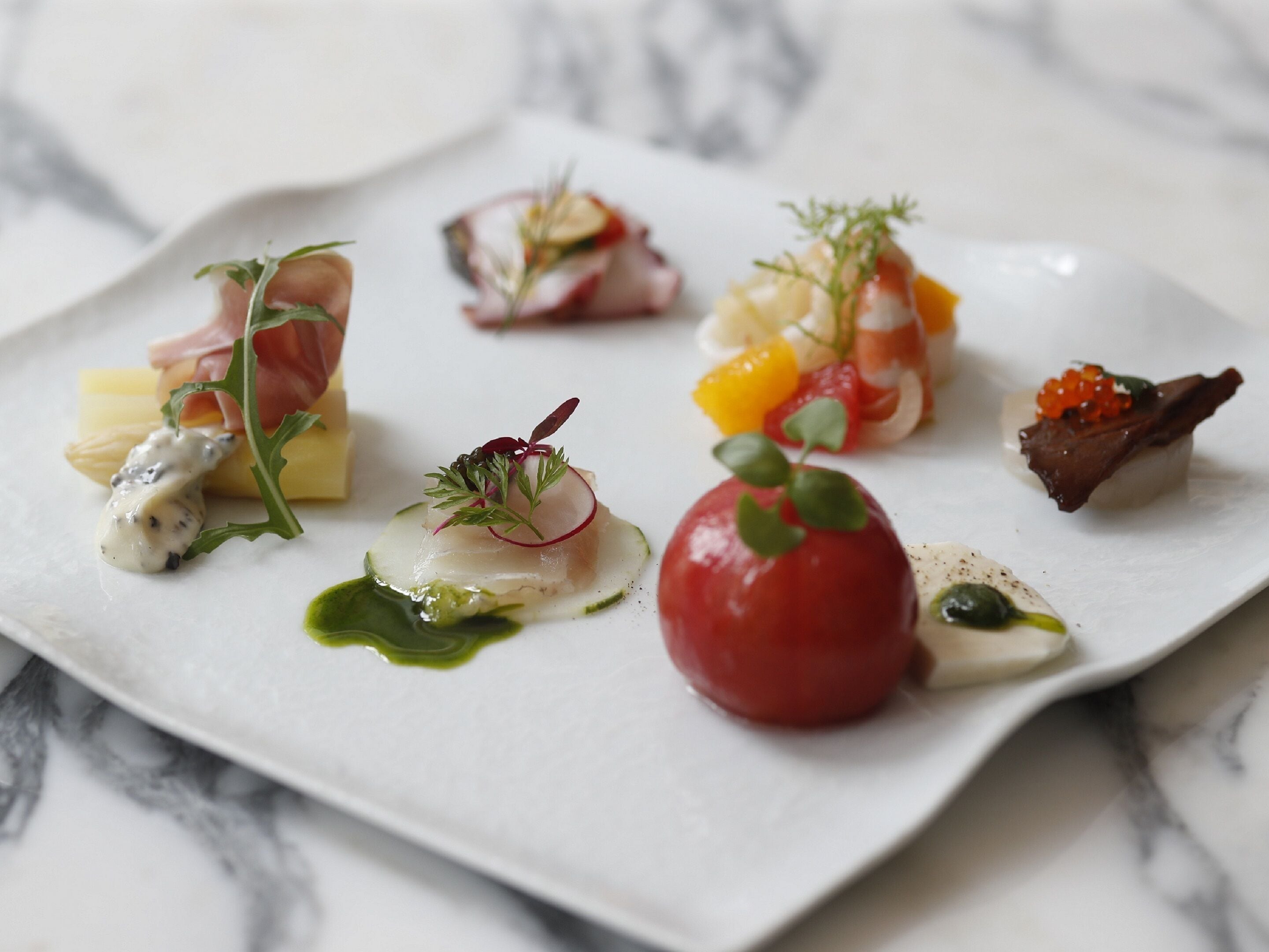 A white plate with various food items, including a red tomato, some fish, and garnishes.