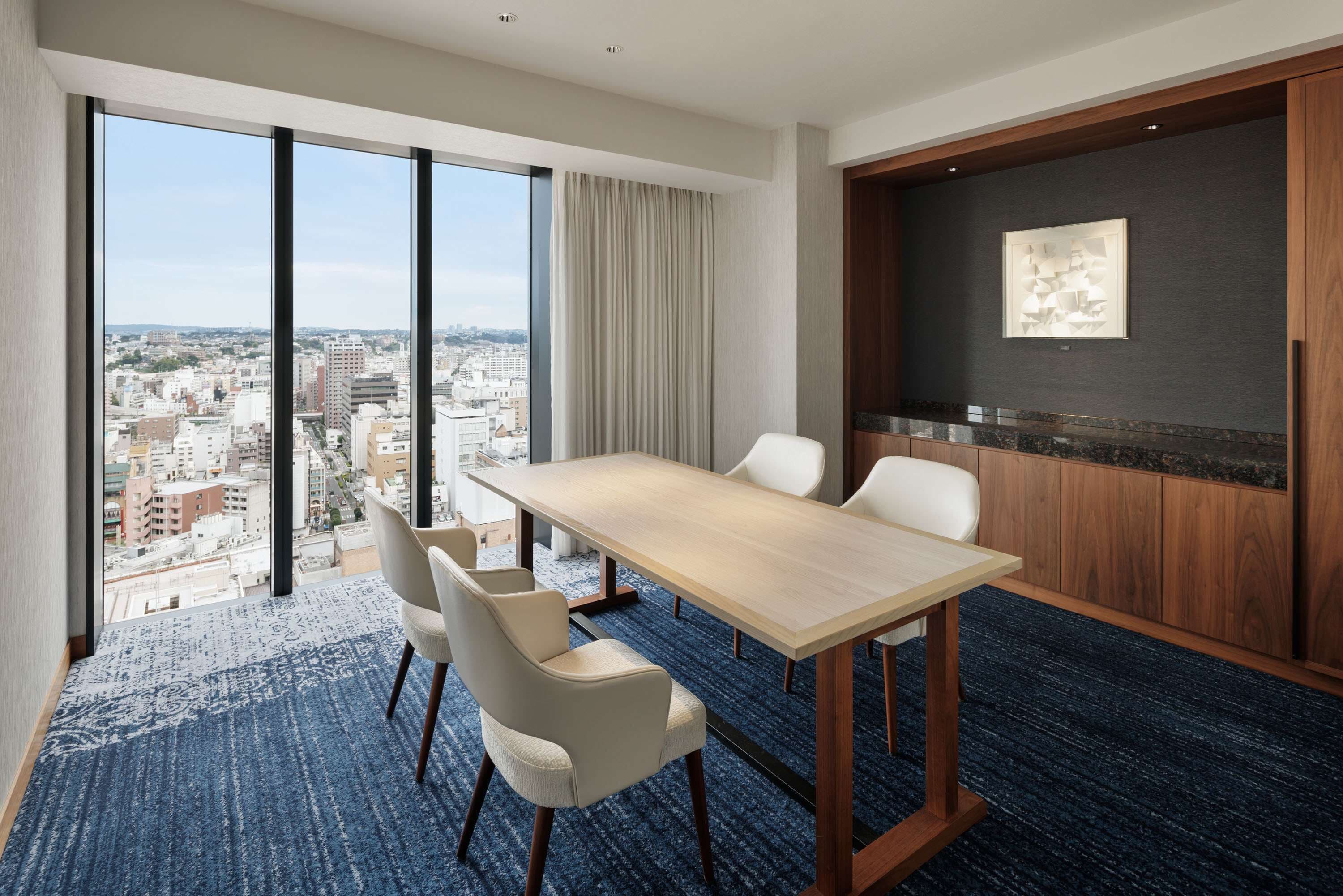 A hotel room with a large window offering a city view, a wooden dining table with chairs, and a dark wood paneling wall.