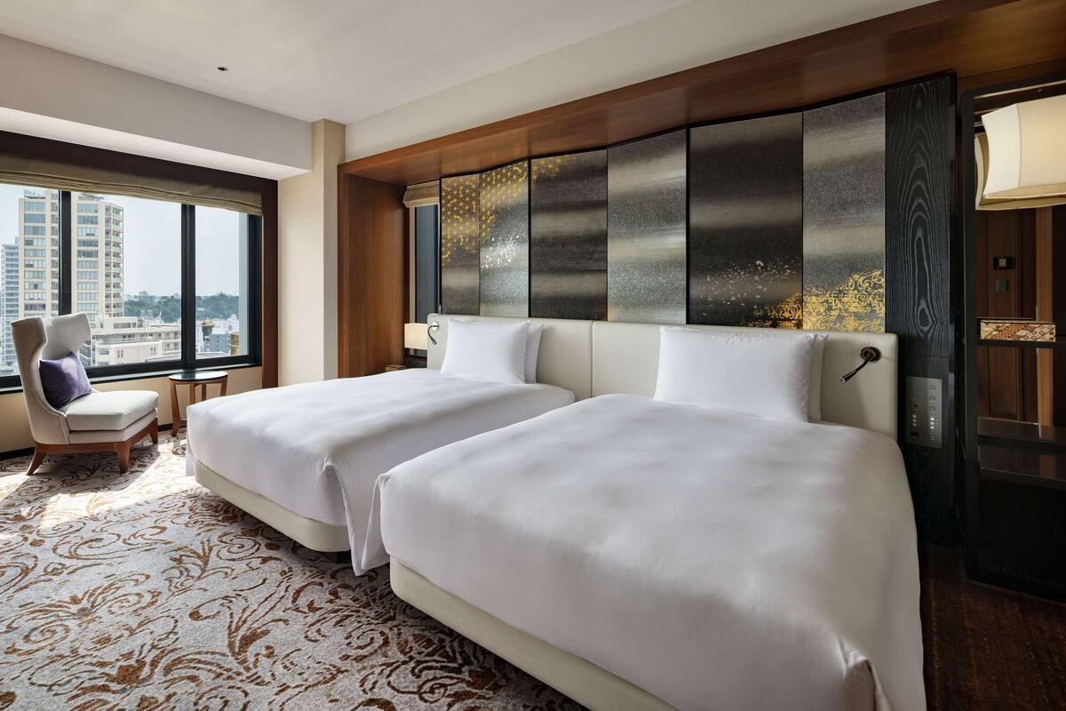 A hotel room with two beds, a large window, a chair, and a decorative headboard.