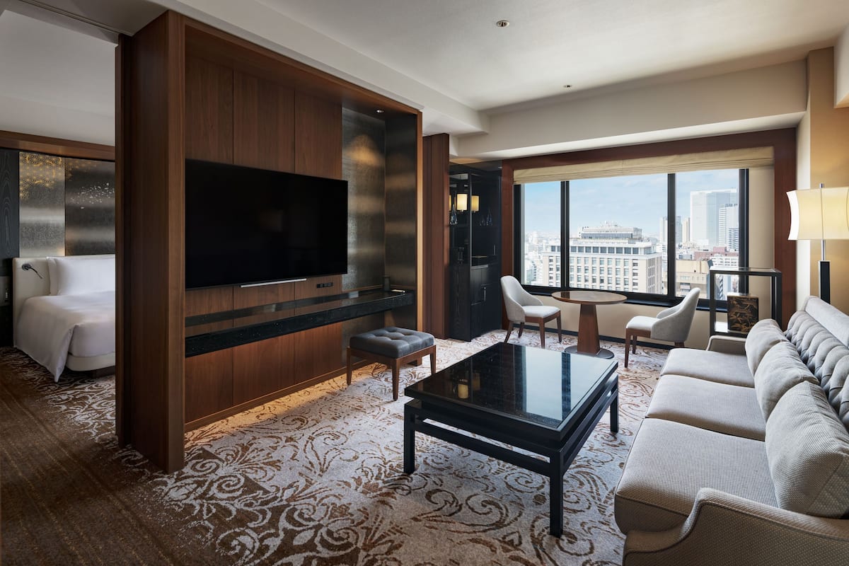 A modern hotel room with a large flat-screen TV, a sofa, a coffee table, and a city view through large windows.