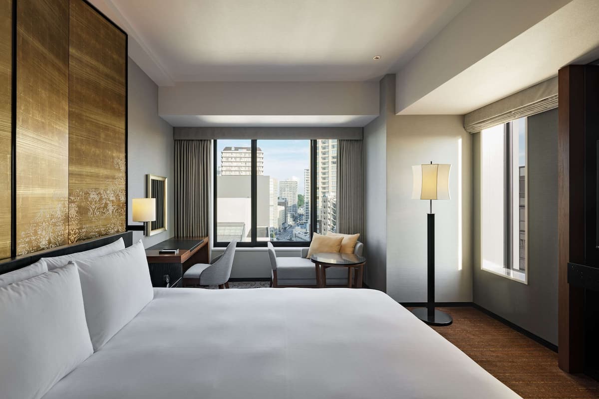 A hotel room with a large bed, a desk, a chair, a floor lamp, and a view of the city through large windows.
