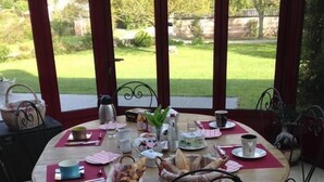 Une table ronde dressée pour un repas, entourée de chaises, avec vue sur un jardin, visible à travers une zone couverte agrémentée d’arcades.