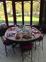 Mesa redonda posta para refeição, com cadeiras, vista para um jardim através de um espaço coberto com arcos.
