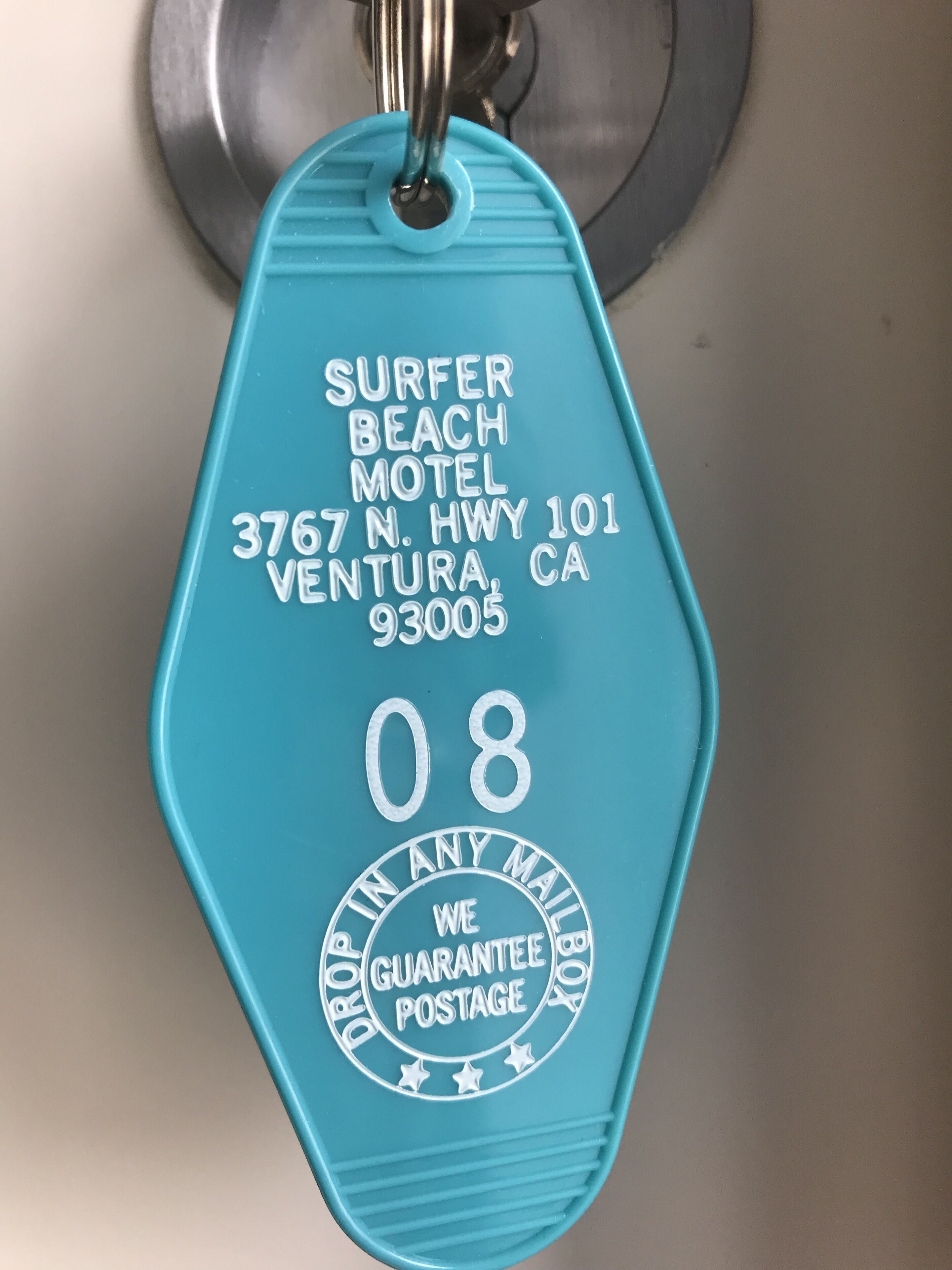 A turquoise key tag with the text "SURFER BEACH MOTEL, 3767 N. HWY 101, VENTURA, CA 93005, 08, POSTAGE GUARANTEE".