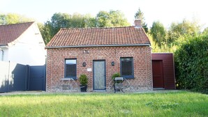 Une petite maison en briques avec un toit en tuiles, une porte d’entrée et un petit jardin avec de l’herbe et une plante en pot.