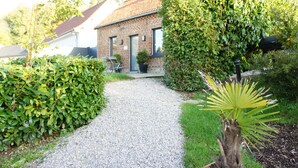 Un chemin de gravier mène à une maison en briques avec une porte verte, entourée d’une végétation luxuriante et d’une plante ressemblant à un palmier.