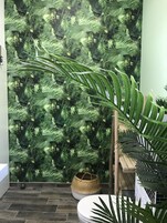 Um banheiro com parede estampada em folhas verdes, um banco de madeira e um vaso com planta.
