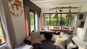A modern living room with a sectional sofa, a dining table, and a large window with a view of greenery. - Family-friendly, cozy accommodation with a beautiful garden plot  (Bruinisse)