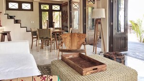 Um quarto espaçoso com teto de madeira, uma cama, uma cadeira de madeira, um banquinho de fibra natural e um espaço de refeições com cadeiras e uma mesa.