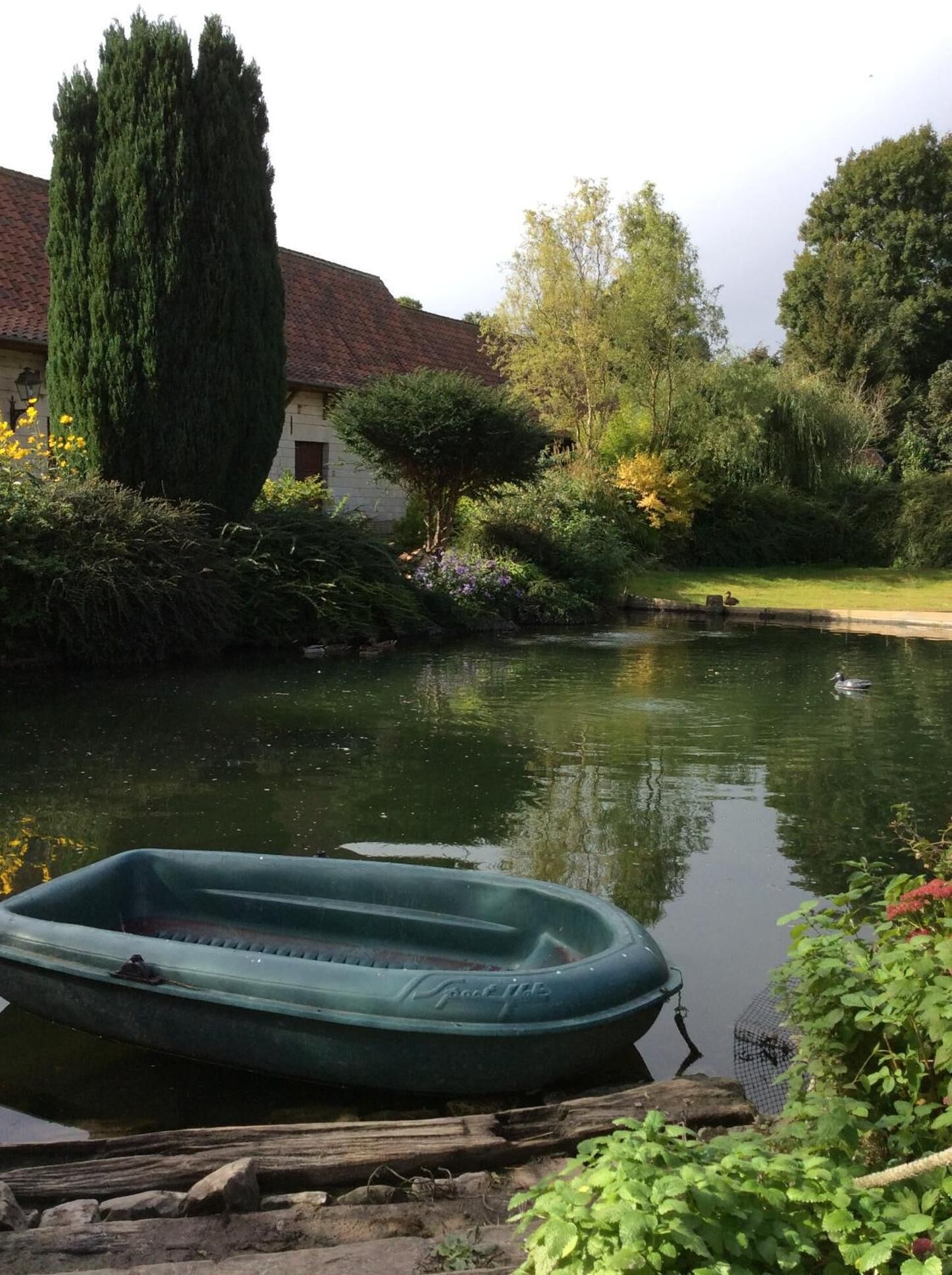 Un río tranquilo con una balsa inflable verde amarrada en la orilla, rodeado de abundante vegetación y un edificio de techo rojo.