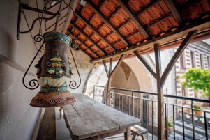 Uma área de estar rústica ao ar livre com uma mesa e bancos de madeira, um candeeiro de metal decorativo e vista para um edifício com arcos ao fundo.