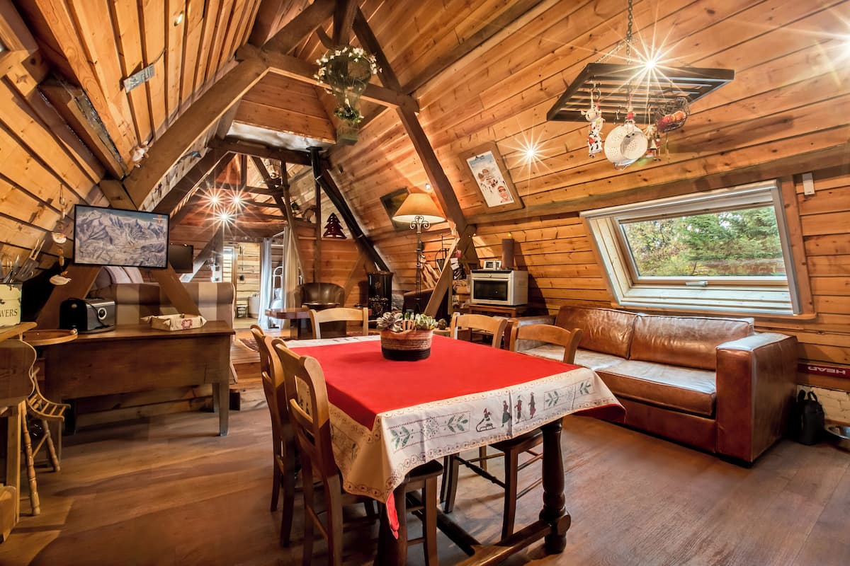 A cozy cabin interior with a dining table, wooden chairs, a sofa, and a large window.
