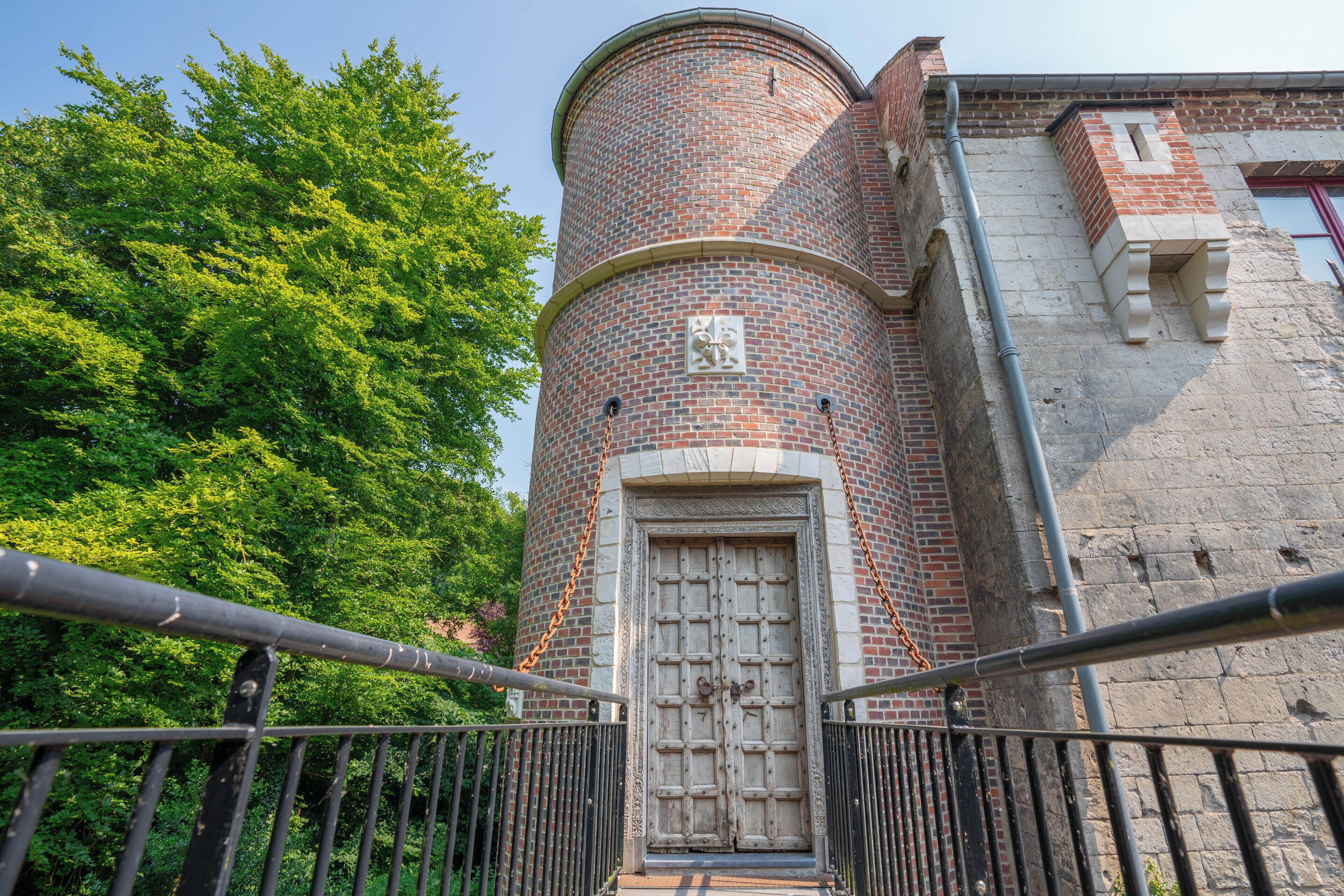 A brick tower with a large wooden door, surrounded by greenery and a metal railing.