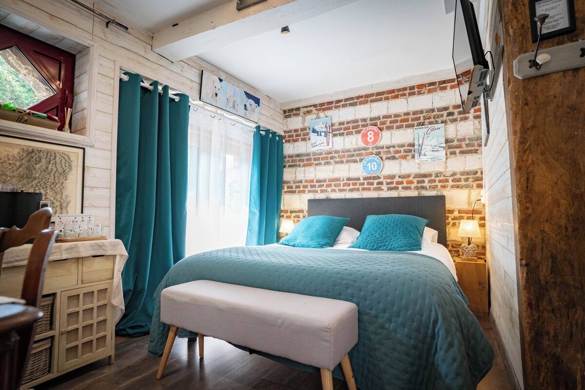 A bedroom with a brick wall, a bed with teal bedding, a wooden bench, a desk with a chair, and a window with curtains.