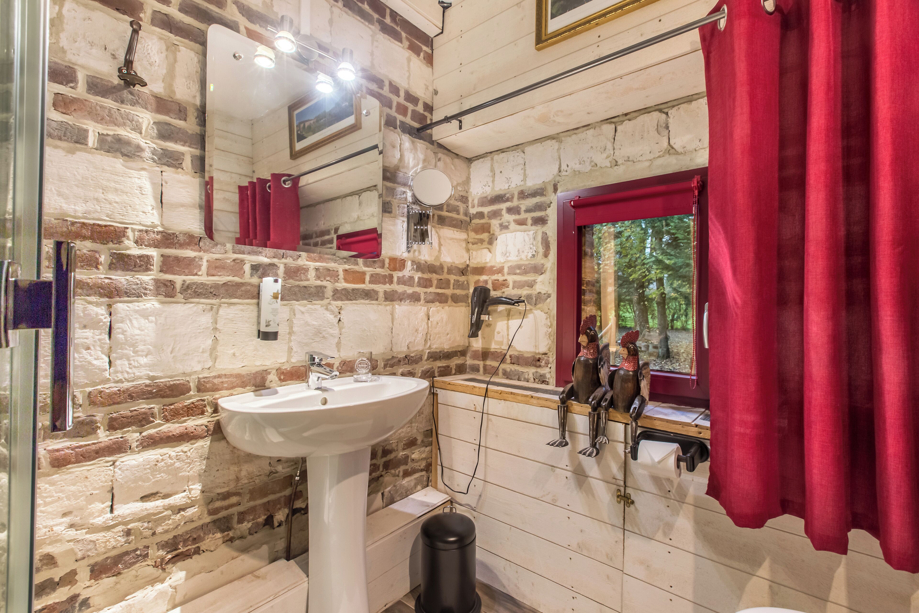 A bathroom with a white sink, red curtains, a window with a view of trees, and a rustic brick wall.