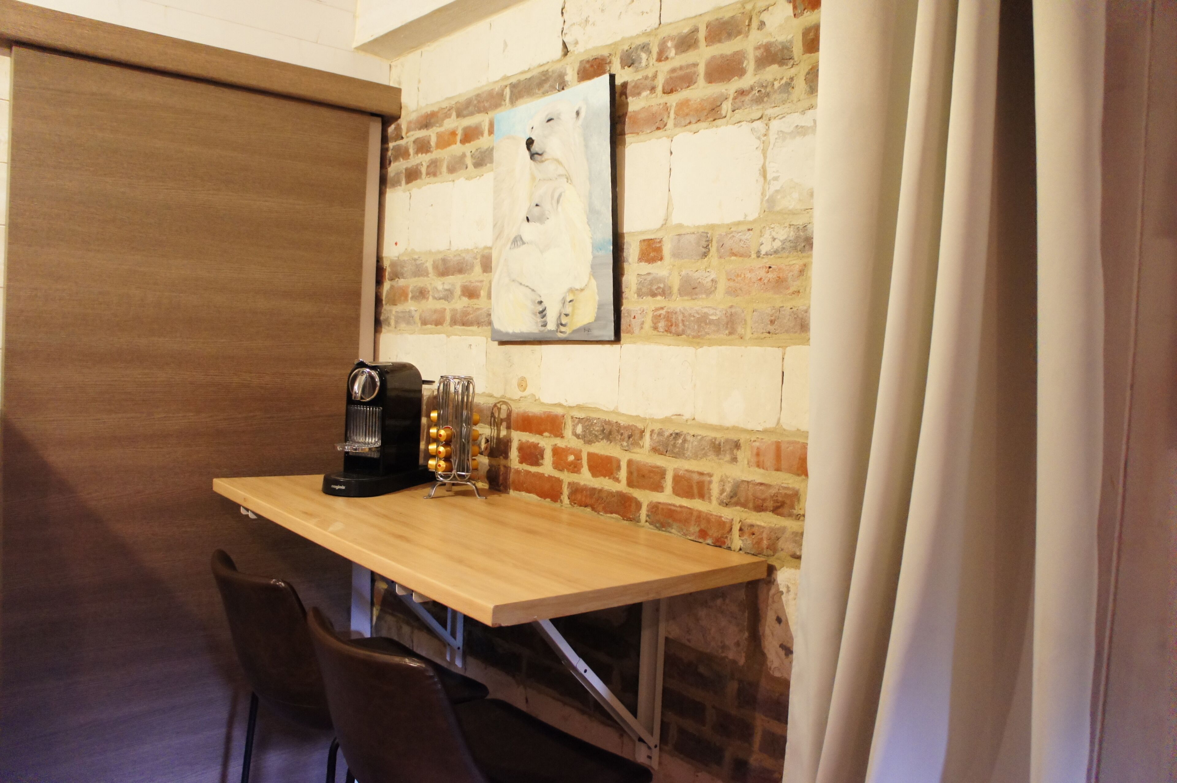A small dining area with a wooden table, two chairs, a coffee machine, and a painting on the brick wall.