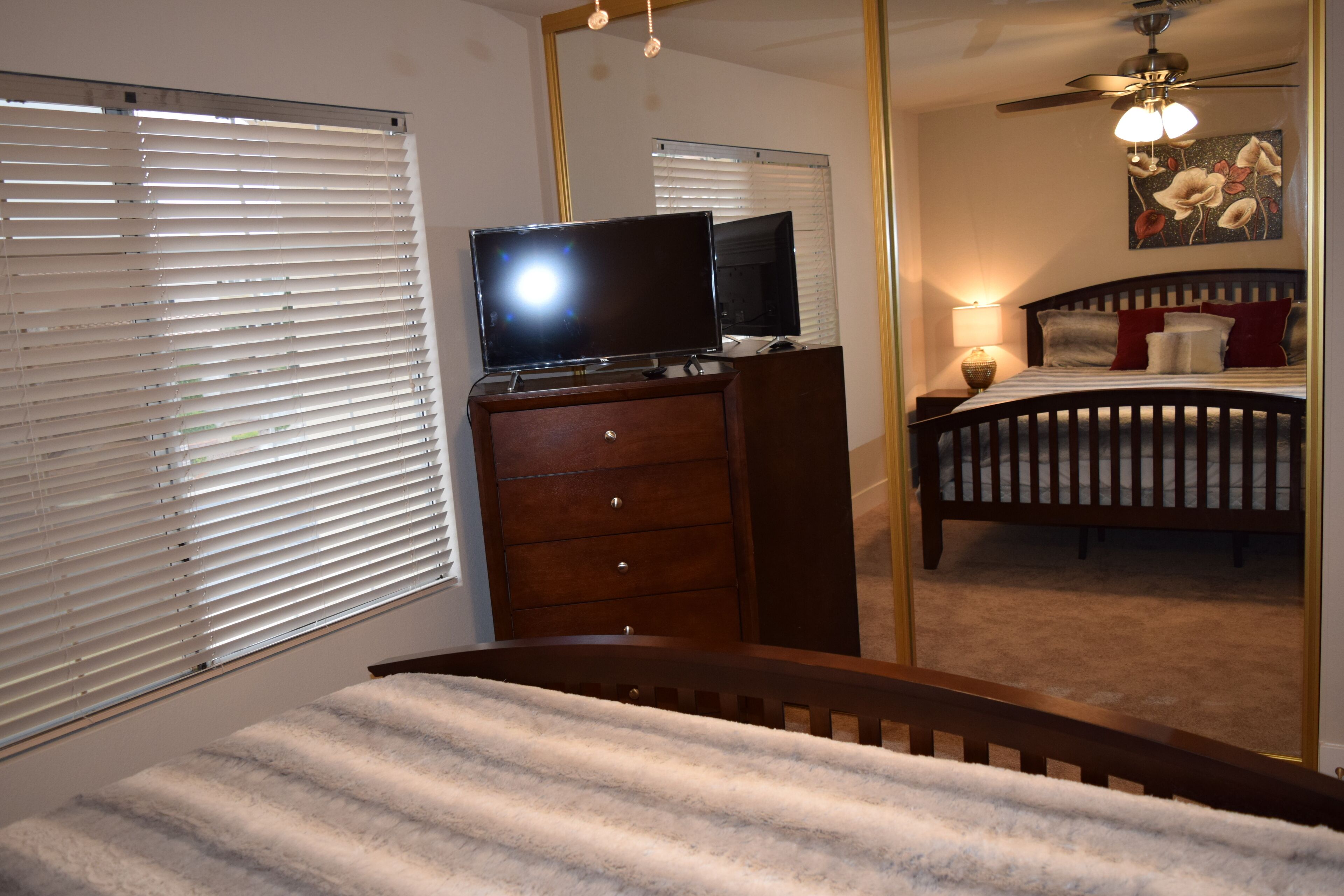 A bedroom with a bed, a dresser, a television on top, a ceiling fan, and a painting on the wall.