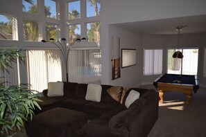 A living room with a brown sofa, a pool table, and a large window with blinds.