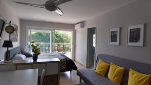 Ein modernes Wohnzimmer mit grauem Sofa, gelben Kissen, Deckenventilator, Bett mit Blick auf einen Balkon und einer Essecke mit Tisch und Stühlen.