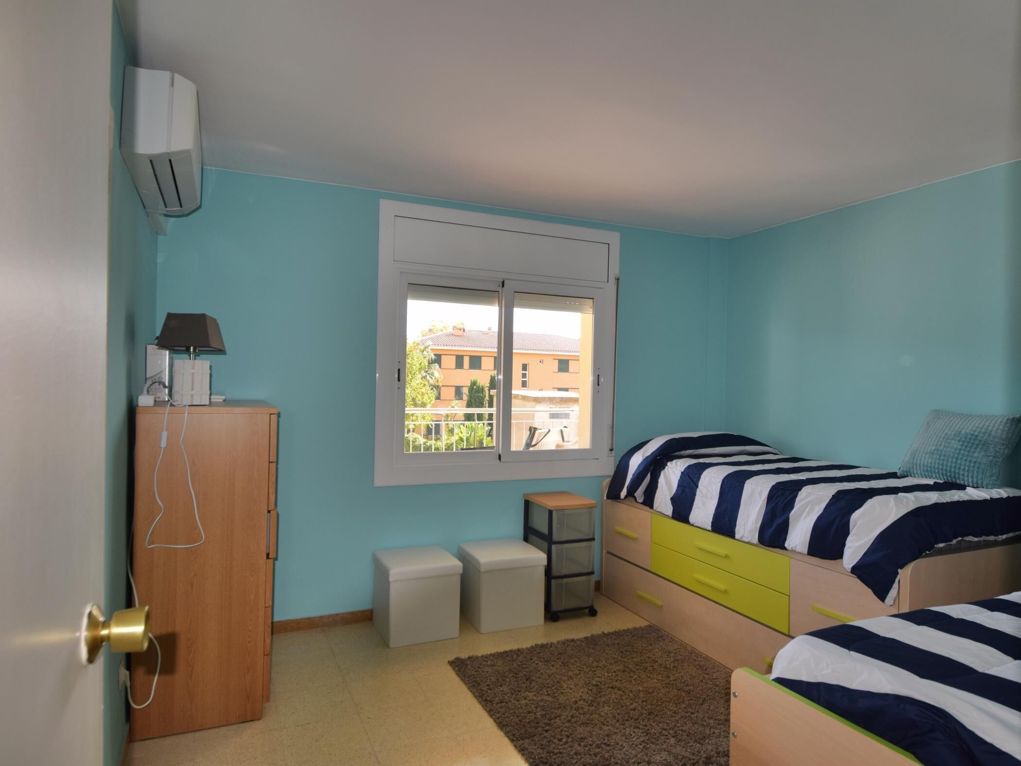 A bedroom with two beds, a desk, a wardrobe, and a window with a view of buildings.