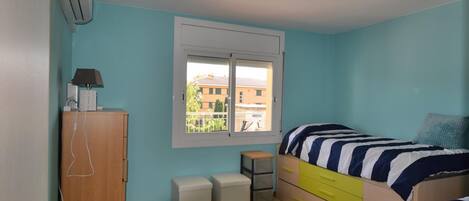A bedroom with two beds, a desk, a wardrobe, and a window with a view of buildings.