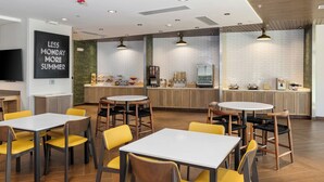 A modern cafe with round tables and yellow chairs, a kitchen area with a coffee machine, and a sign on the wall.
