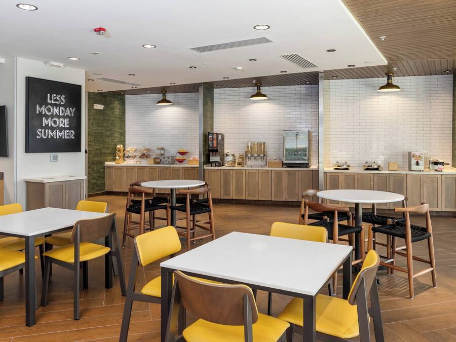 A modern cafe with round tables and yellow chairs, a kitchen area with a coffee machine, and a sign on the wall.