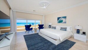 Quarto com cama grande, escrivaninha e cadeira. Janelas oferecem vista para a praia e oceano.