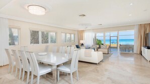 Uma sala ampla com uma mesa de jantar, cadeiras brancas, um sofá e uma televisão. Janelas grandes oferecem vista para o mar.