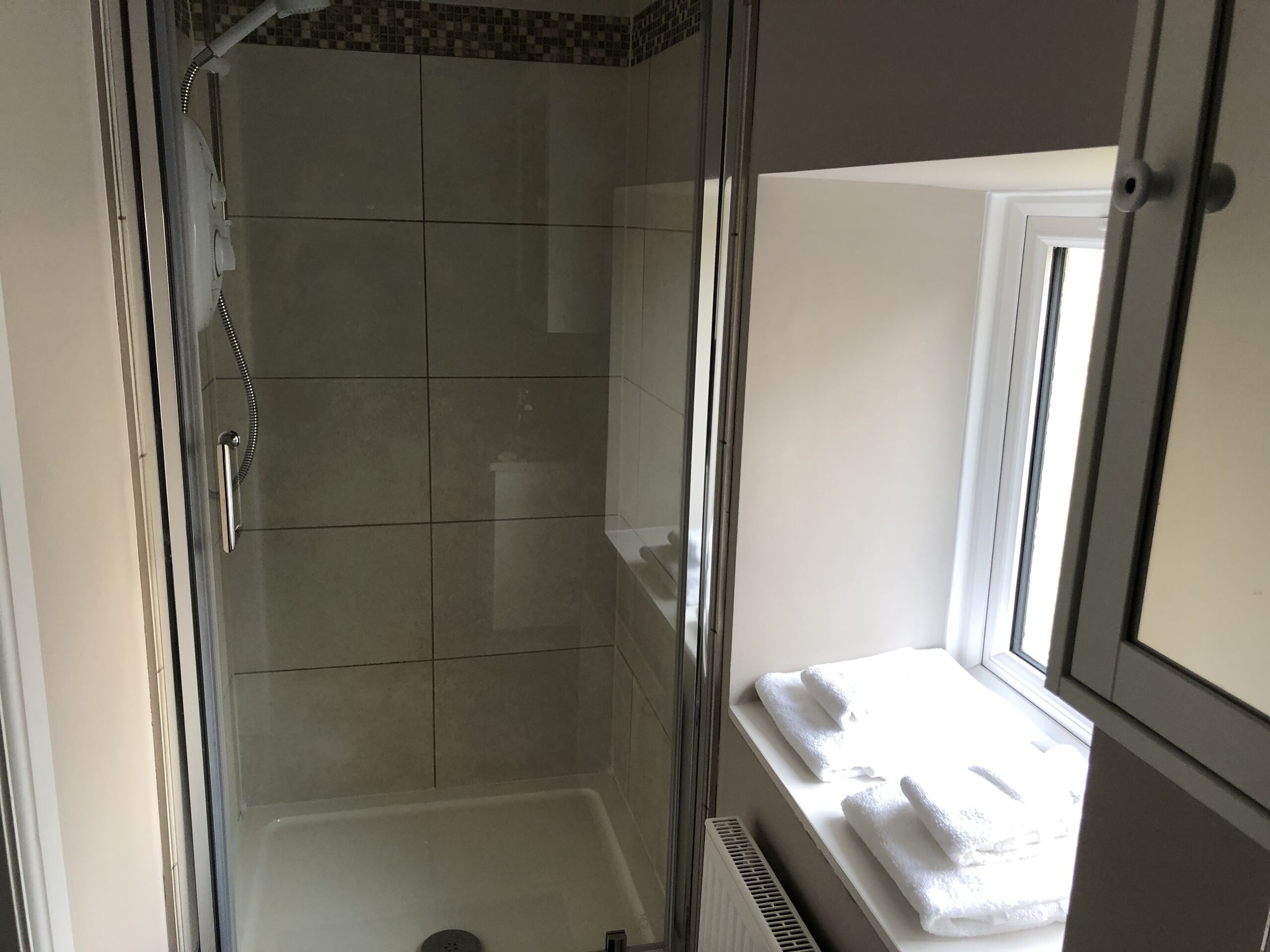 A bathroom with a glass shower enclosure, tiled walls, a window with a towel rack, and a radiator.