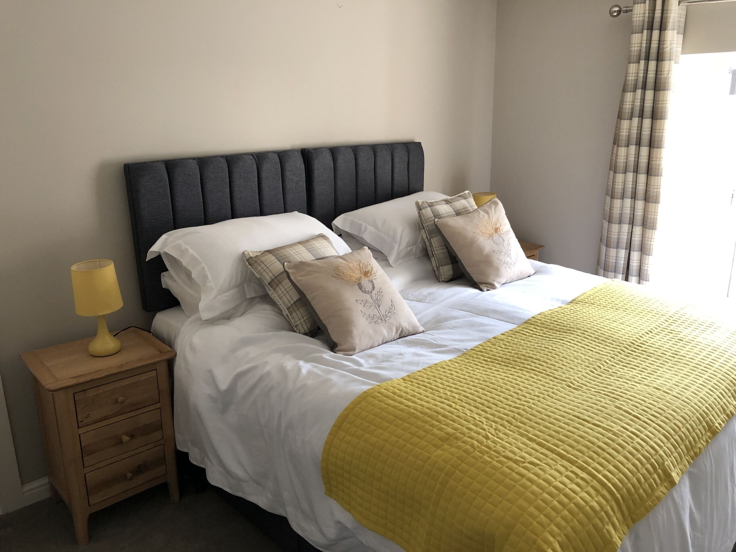 Un lit bien fait, avec du linge de lit blanc et une couverture jaune matelassée, flanqué de deux tables de chevet avec des lampes, dans une pièce dont la tête de lit est sombre et les murs sont de couleur unie.